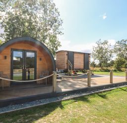 An outdoor wooden pod with glass doors and chairs on a deck next to a small wooden cabin on stilts at Hawkstone Kenstone near Hodnet