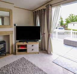 A living room with a television on a wooden stand next to curtains and a door leading to a deck with seating at Osprey 6 in Kiln Park Tenby