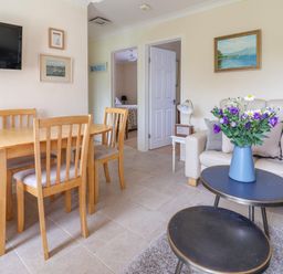 A dining table with four wooden chairs a wall mounted TV a sofa with cushions and a coffee table with a vase of purple flowers at Seaweed in Weston near Sidmouth