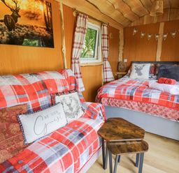 A small room with a bed, a sofa covered in red and gray plaid blankets, a small wooden table, and a window with plaid curtains at Shepherd's Cwtch in Cilycwm near Llandovery