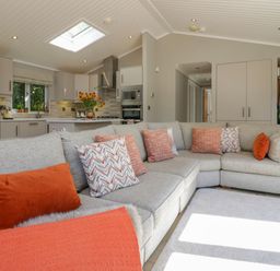A living room with a large gray sectional sofa with orange and patterned cushions and a kitchen with gray cabinets and appliances at Waterside Lodge 55A Bowness White Cross Bay near Windermere