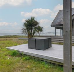 An outdoor wooden deck with rattan furniture near a grassy area a palm tree and the sea at Dunes in Rhosneigr