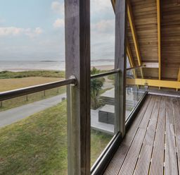A balcony with wooden floor and glass railing overlooking a grassy area and beach at Dunes in Rhosneigr