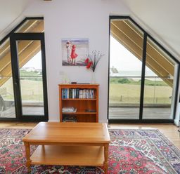 A living room with a wooden coffee table a patterned rug a bookcase and double glass doors overlooking a grassy area and the sea at Dunes in Rhosneigr