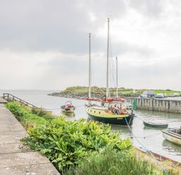A harbor scene with sailboats and small boats tied at the dock by a walkway with plants and cars parked on the road at Lazey Cottage in Haverigg