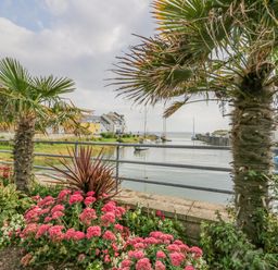 A waterfront view with palm trees and pink flowers along a railing near boats and houses at Lazey Cottage in Haverigg