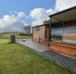 A wooden lodge with a covered porch and glass railing next to a grassy lawn and fenced field at Math Lodge 3 in Llanelidan near Corwen