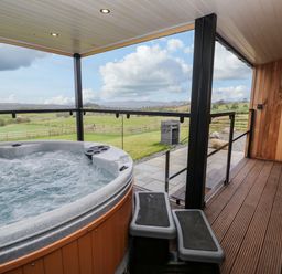 An outdoor covered patio with a hot tub wooden deck stairs glass railing and a grassy field beyond at Math Lodge 3 in Llanelidan near Corwen