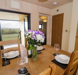 A dining table set with plates, bowls, wine glasses, a vase with flowers, and a bottle in a room with wooden walls and doors at Math Lodge 3 in Llanelidan near Corwen
