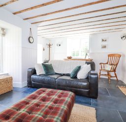A living room with a black sofa with pillows a plaid ottoman a wooden chair and window at Hilldene in Treknow