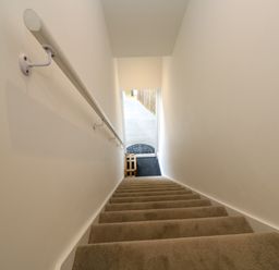 A carpeted staircase with a handrail on the left leading down to a wooden stool on a dark floor at Amelia's Point in St Ives