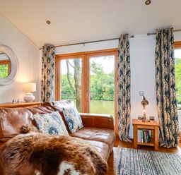 A living room with a leather sofa and patterned curtains by large glass doors overlooking a river at 10 Water's Edge in Lanreath