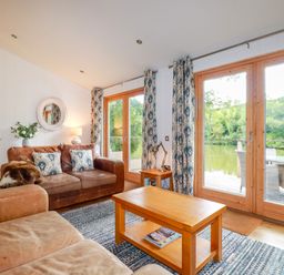 A living room with leather sofas wooden coffee table and large glass doors overlooking water at 10 Water's Edge in Lanreath