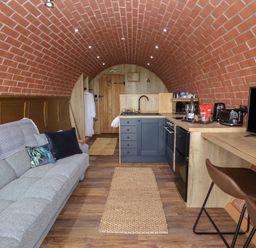 A narrow living space with a grey sofa and cushions a kitchen area with blue cabinets and a wooden countertop a wooden bar with two brown stools and a TV on the right Rhosydd Cyffylliog near Ruthin