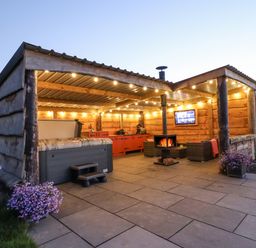 An outdoor covered patio with a hot tub chairs a wood stove string lights and a TV at Cae Bach in Cyffylliog near Ruthin