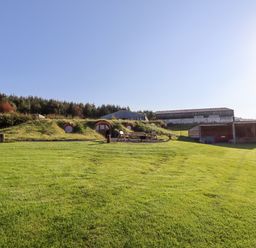 A grassy outdoor area with a hobbit-style house, wooden chairs around a fire pit and farm buildings in the background at Weirglodd in Ruthin