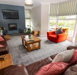 A living room with brown leather sofas a red armchair wooden tables a TV mounted on a blue wall and windows with striped blinds at Morva in New Quay