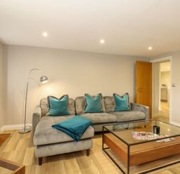 A living room with a gray sectional sofa with teal pillows and blanket a metal floor lamp and a glass coffee table at Daisys Place in South Creake near Little Walsingham