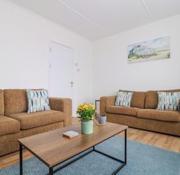 A living room with two brown sofas, a wooden coffee table, a potted plant, and a wall painting at Small Hillman in Perranporth