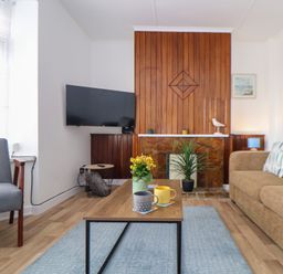 A living room with a wooden coffee table with mugs and a flower pot a TV mounted on the wall and a plant by a fireplace at Small Hillman in Perranporth