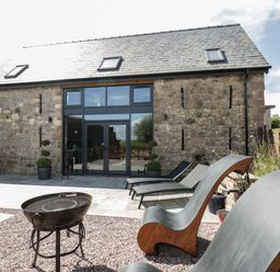An outdoor patio area with modern lounge chairs a barbecue grill and a stone building with large glass doors at The Hay Barn in Huntley