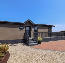 An exterior view of a single-story wooden lodge with stairs and a railing at Hazelwood 2 in Pinewood near Lyme Regis
