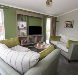 A living room with two sofas, a wooden coffee table with cups, a wall-mounted shelving unit with a TV and decorative items at Hazelwood 2 Pinewood near Lyme Regis