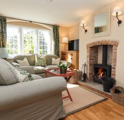 A living room with a sofa, armchair, coffee table, fireplace, wooden floor, and window at The Alms House East in Forthampton near Tewkesbury
