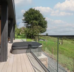 An outdoor wooden deck with a covered hot tub and glass railing overlooking a green field at The Lookout in Longridge