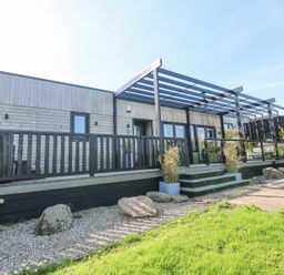 A wooden terrace with glass roof and stairs leading to a pathway with rocks and grass at Bay View in Portreath