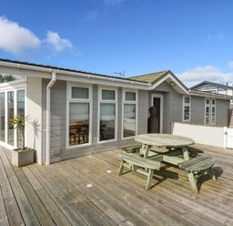 An outdoor wooden deck with a round picnic table and benches next to a building with multiple windows and a glass door at The Warren M45 in Abersoch