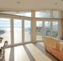 A living area with wooden dining chairs a sofa and large glass doors opening to a deck with ocean view at The Warren M45 in Abersoch