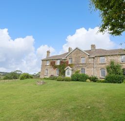 A stone house with chimneys surrounded by shrubs and grass under a blue sky at Ivy Cottage in Colerne