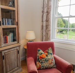 A red armchair with a patterned cushion next to a wooden cabinet with books and DVDs and a table lamp by a window with floral curtains at Ivy Cottage in Colerne