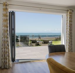 A dining area with a round wooden table and chairs overlooking a patio with lounge chairs and an ocean view at Melin Y Plas in Llanaber