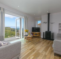 A living room with two grey sofas, a round wooden coffee table, a television on a wooden stand, a wood-burning stove, and open doors leading to a balcony with a view of green hills and the sea at Rowena in Mullion