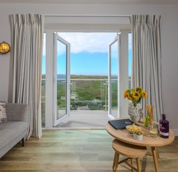 A living room with a grey sofa and open double doors leading to a balcony overlooking a green landscape at Rowena in Mullion