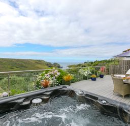 An outdoor deck with a hot tub a dining table with chairs and an umbrella overlooking hills and the sea at Morwenna in Mullion