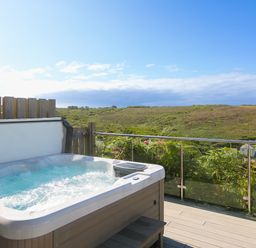 A bubbling hot tub on a deck overlooking green hills with a glass railing at Verity in Mullion