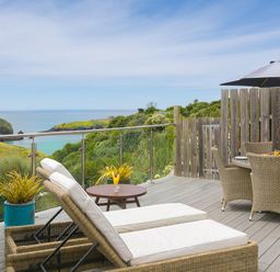 An outdoor deck with wicker sun loungers and a dining table under an umbrella overlooking coastal cliffs and the sea at Daphne in Mullion