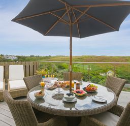 A round glass outdoor dining table with wicker chairs and a large umbrella on a deck with breakfast items and a view of green hills and houses at Daphne in Mullion