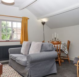 A living room with a grey sofa and two cushions a wooden dining table with chairs and a candelabra by a window with yellow curtains at Hole Farm Stables in Bickington near Ashburton
