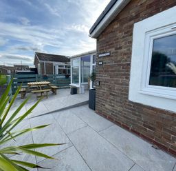 A patio area with a wooden picnic table and benches a potted plant and a brick house with a window at Villa Rose Beadnell near Seahouses
