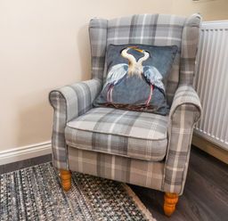 A plaid upholstered armchair with wooden legs and a cushion with two birds on it in a room with a carpet and radiator at Ramson in Saltburn-By-The-Sea