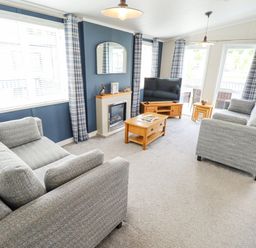 A living room with two grey sofas a wooden coffee table a television on a wooden stand a fireplace and windows with plaid curtains at Sunset Lodge in Woodhall Spa