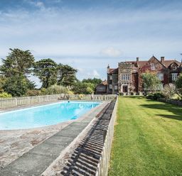 An outdoor swimming pool with a stone patio and a large stone house with greenery in the background at Alston Hall in Battisborough Cross