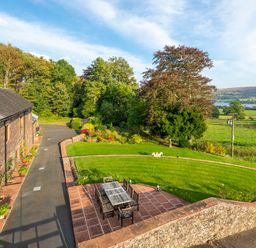 An outdoor patio with a table and chairs next to a stone building and a curved lawn with trees and fields in the background at Bank Barn Wreay in Watermillock