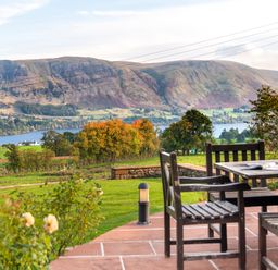 An outdoor patio with wooden chairs and a table overlooking a grassy field with trees a lake and mountains at Bank Barn Wreay in Watermillock