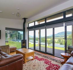 A living room with brown leather sofas a coffee table a wood stove and large windows overlooking a green landscape at 6 Strathtay Lodges in Aberfeldy