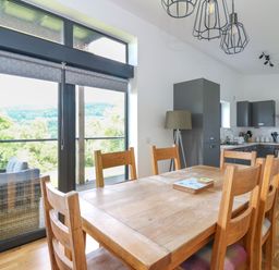 A dining area with wooden table and chairs next to large windows overlooking trees and a kitchen with grey cabinets at 6 Strathtay Lodges in Aberfeldy
